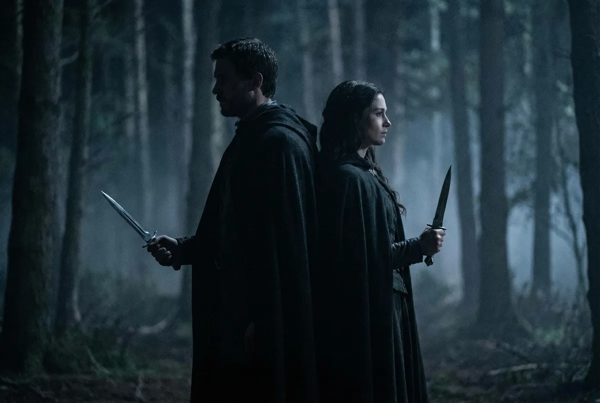 Two cloaked characters standing back to back with daggers in a misty forest, illustrating romantasy character and love interest tension